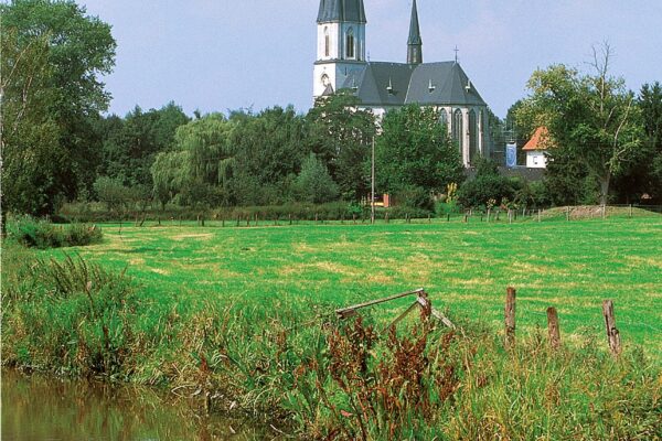Abbildung einer Kirche aus weiter Entfernung mit Wiese und Bach im Bild