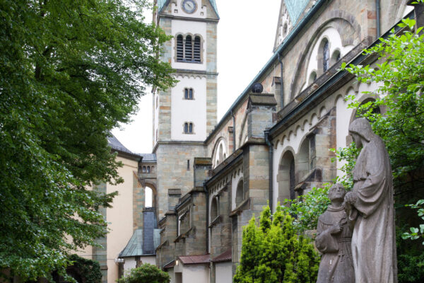 Aussenansicht Kirche mit Garten im Vordergrund und den beiden Türmen im Hintergrund über ein Haus hinausragend